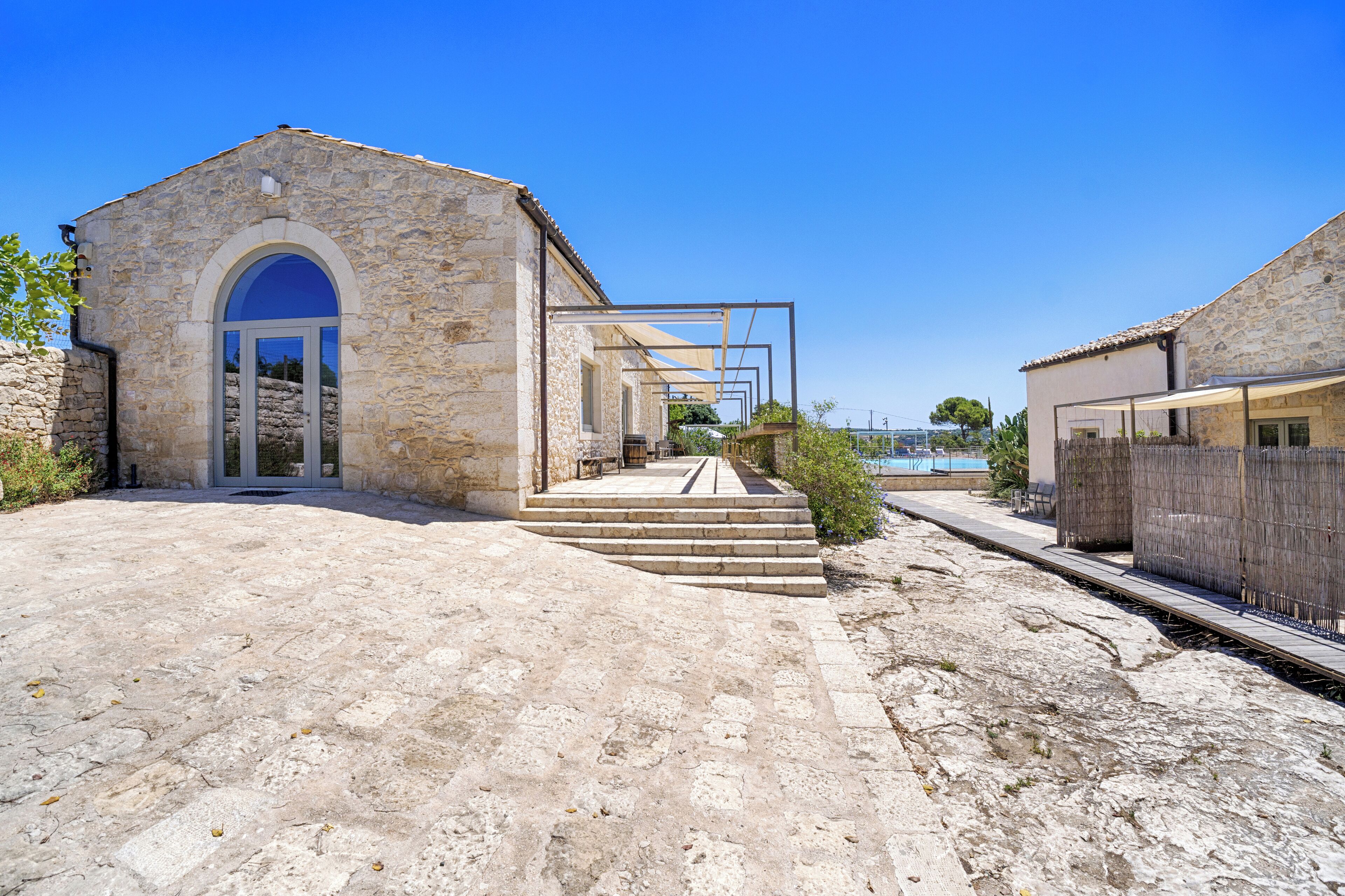 A stone building with a large arched doorway, steps leading to a covered area, and a pool in the background.