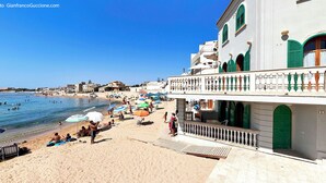 A beachside building with a balcony overlooking the sea, a sandy beach with umbrellas, and people enjoying the water.