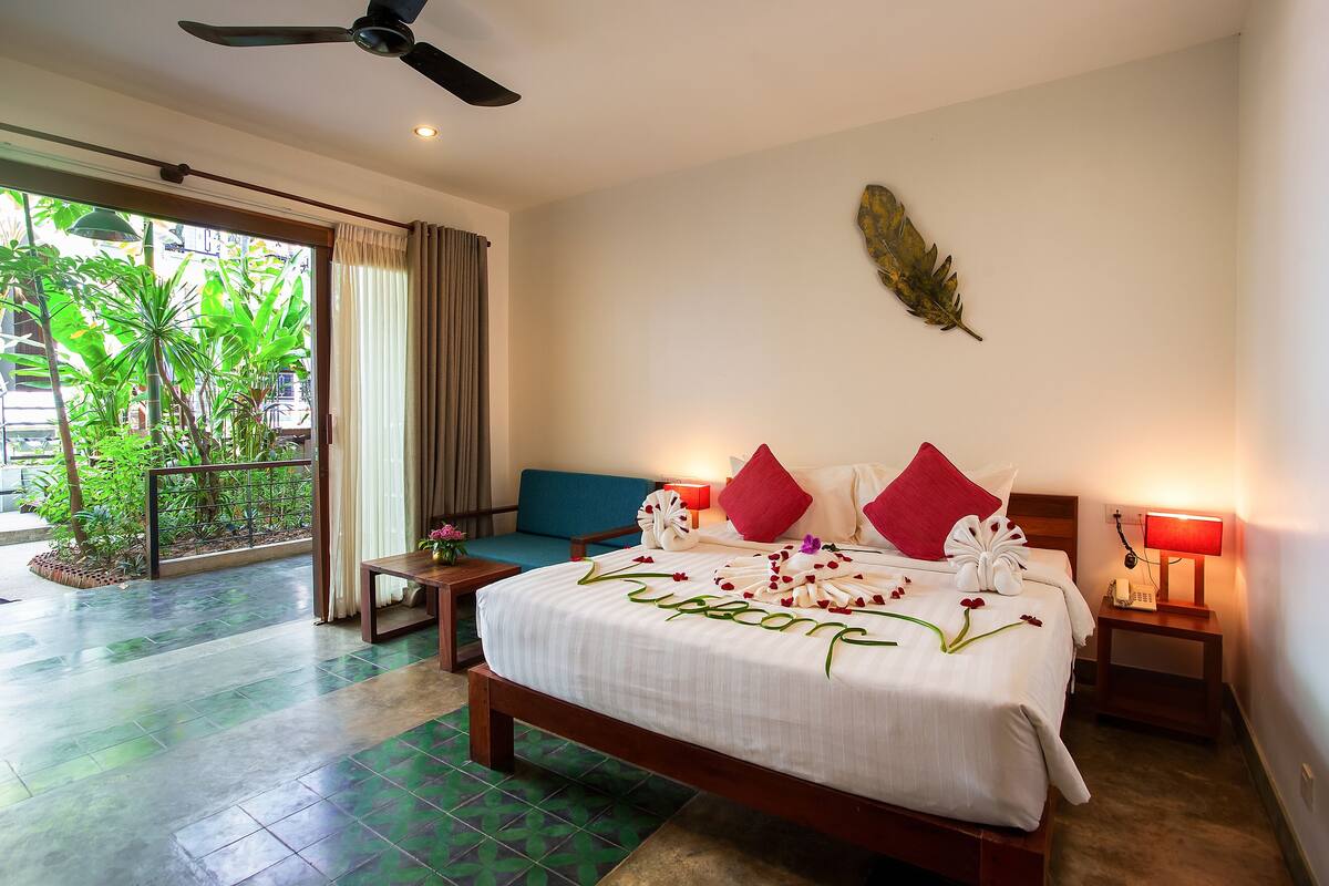 A hotel room with a bed, a desk, a chair, a balcony with plants, and a ceiling fan.
