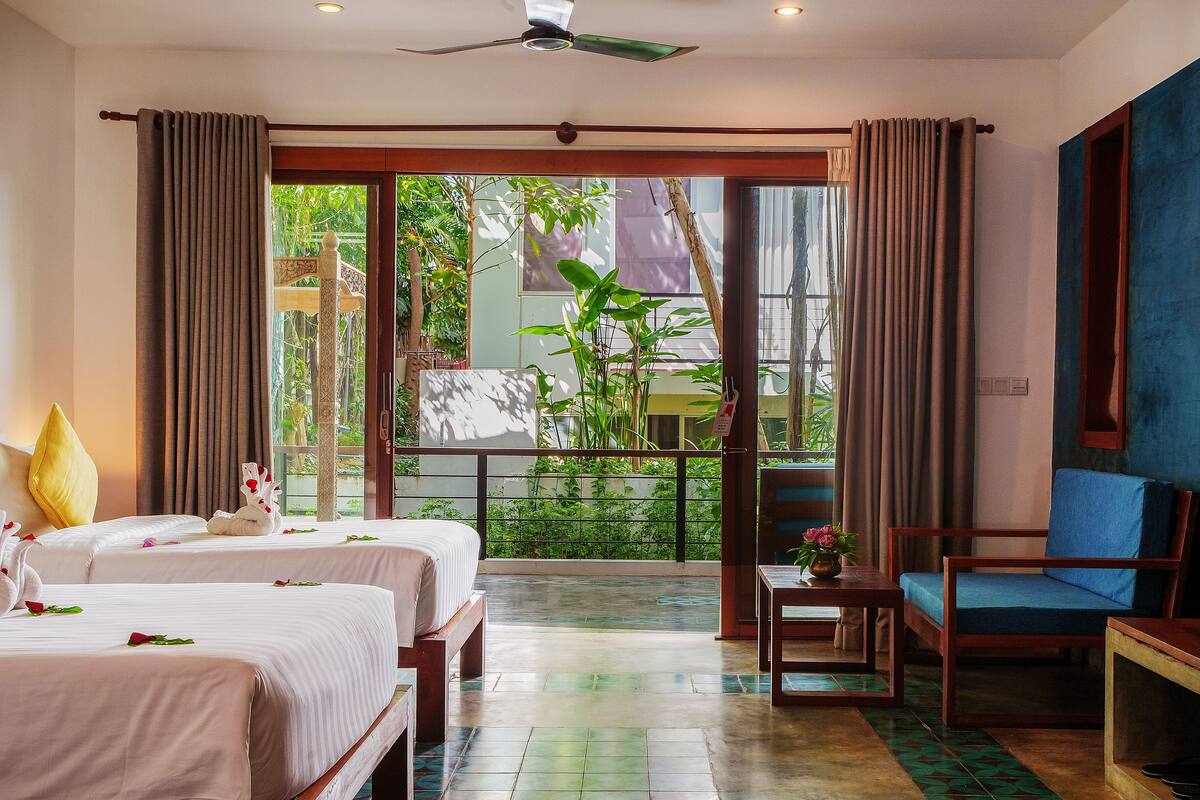 A hotel room with two beds, a balcony with a view of greenery, a ceiling fan, and a blue wall.