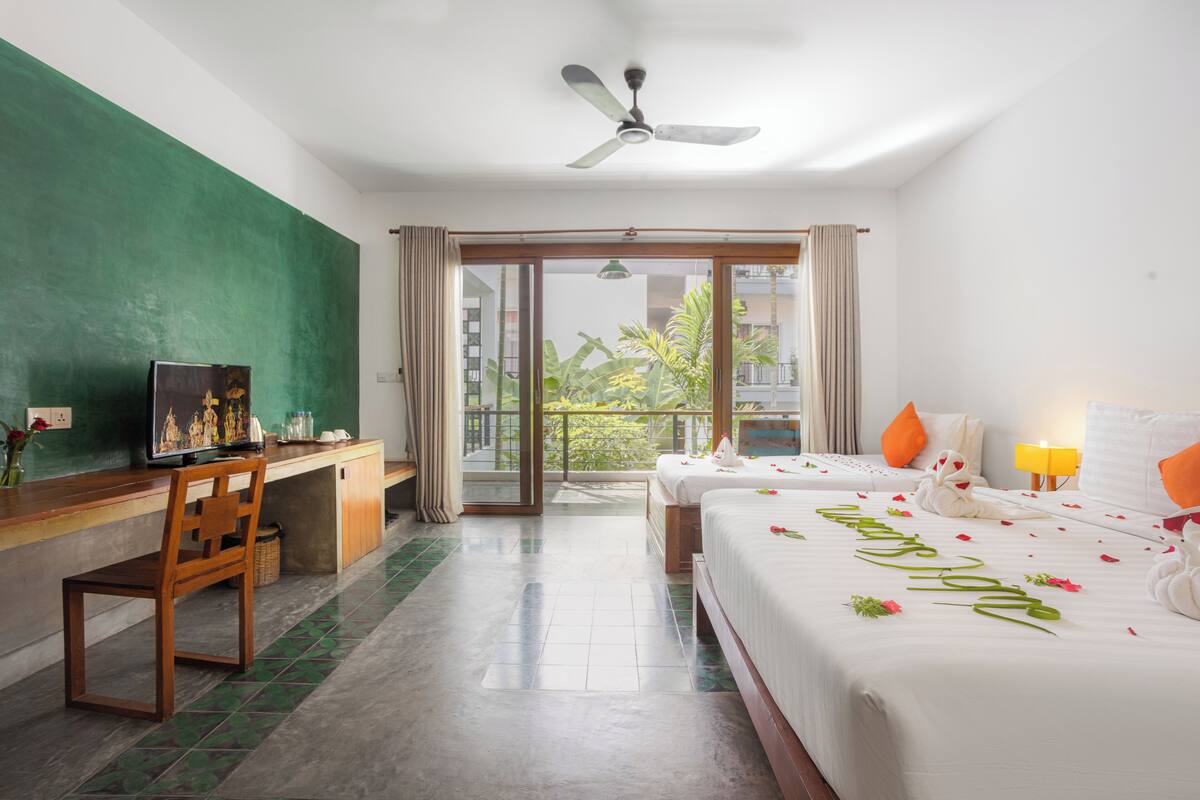 A hotel room with a desk, two beds, a ceiling fan, and a balcony with a view.
