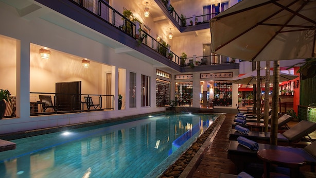 Un área de piscina interior con varios niveles, sillones de descanso y sombrillas, rodeada de paredes con estanterías integradas y plantas en macetas.