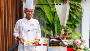 Un chef con uniforme blanco y logotipos, sosteniendo un plato de comida, de pie junto a una estación de cocina con un wok y varios platos.