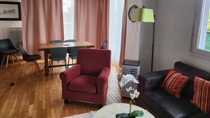 A living room with a wooden dining table, chairs, a red armchair, a black sofa with striped pillows, a floor lamp, and a potted orchid.