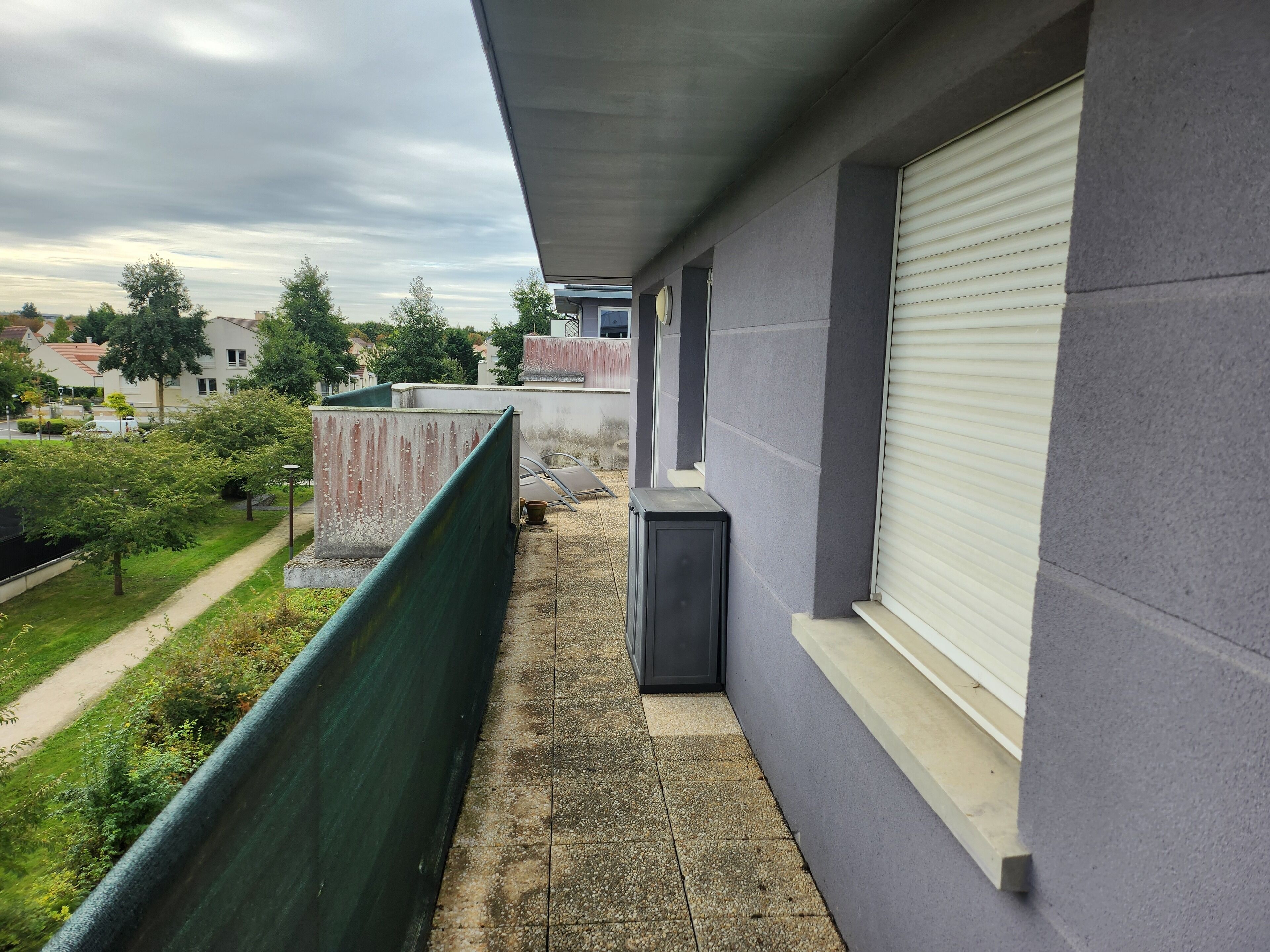 Een smal balkon met een groene veiligheidsbarrière, een raam met gesloten zonwering, en uitzicht op een woonwijk met bomen en huizen.