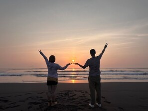 Two people on a beach at sunset, making a heart shape with their hands.