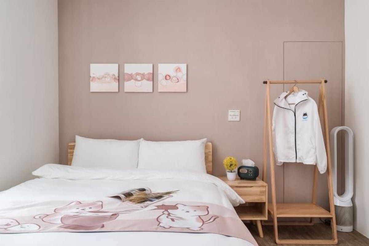 A neatly made bed with a white comforter and a pink blanket featuring cartoon characters. There are three framed artworks on the wall above the bed. A wooden drying rack with a white garment hanging on it is to the right of the bed.
