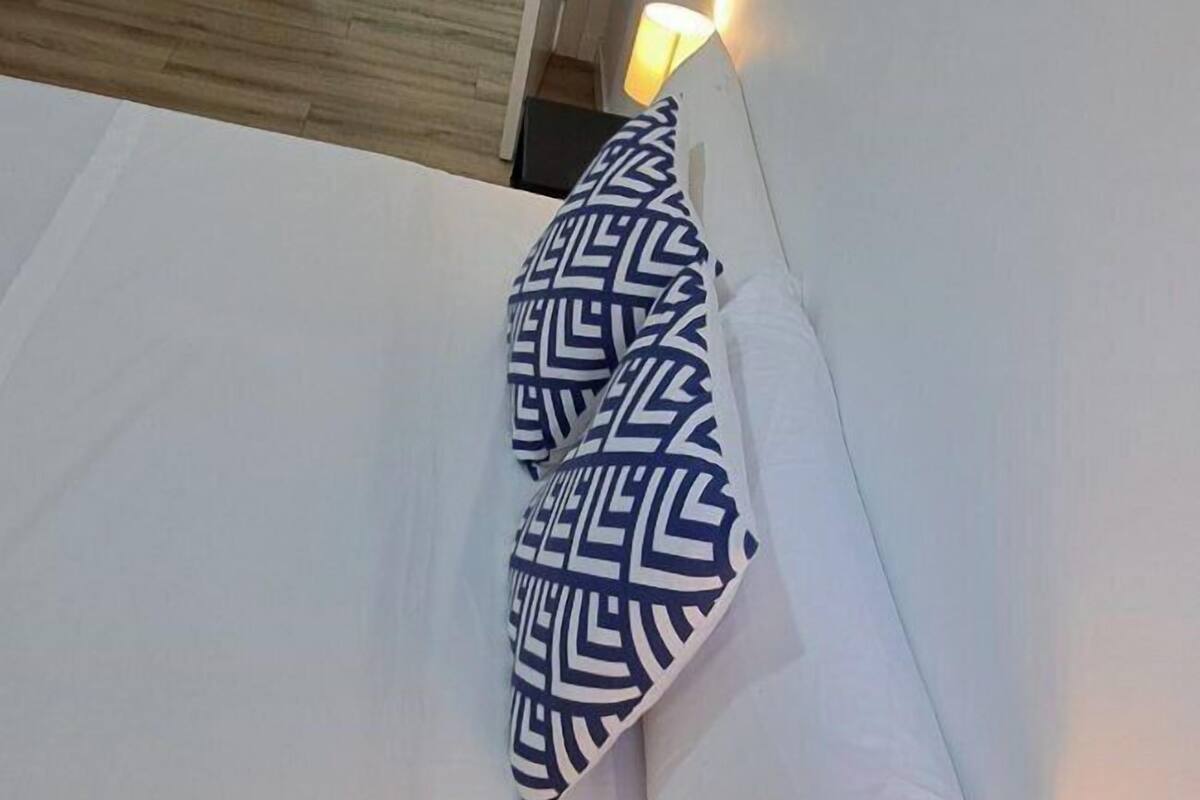 Un lit avec une literie blanche et un oreiller à motifs, une lampe de chevet et une porte donnant sur une autre pièce.