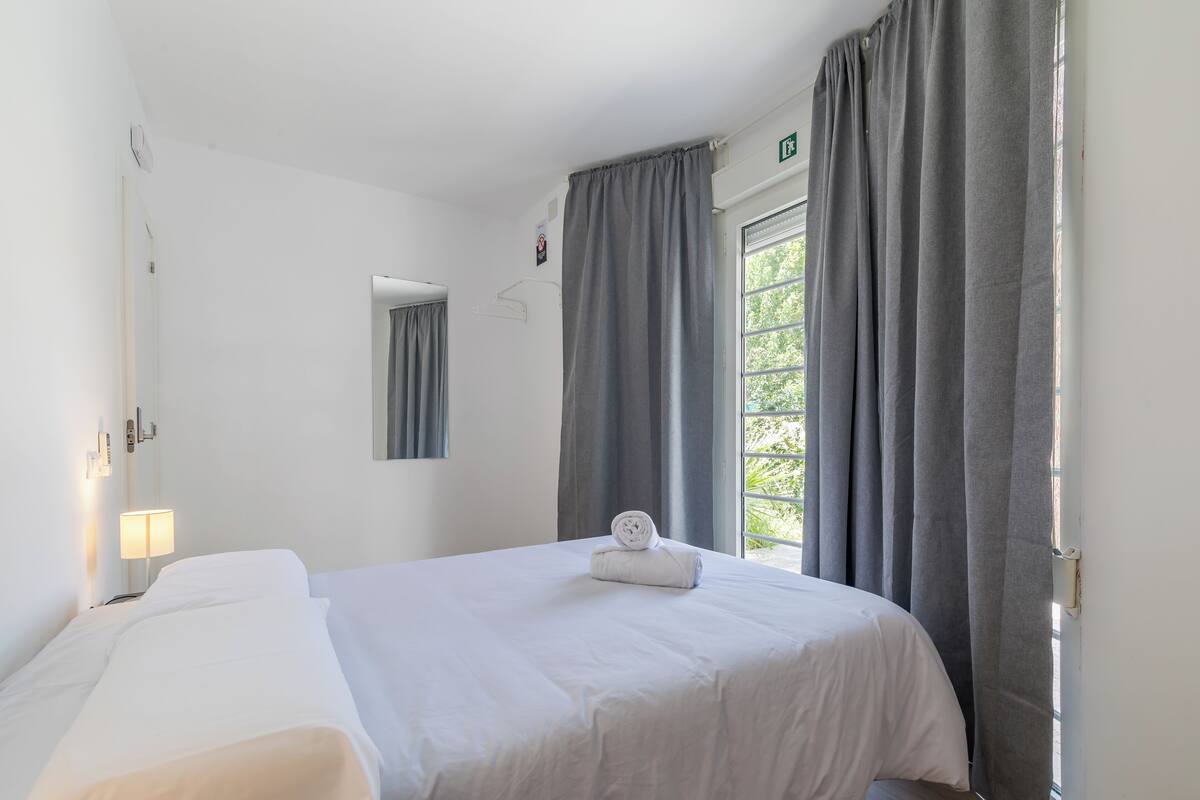 Une chambre d’hôtel avec un lit, une table de chevet, une lampe, une fenêtre avec des rideaux et une vue sur la verdure.