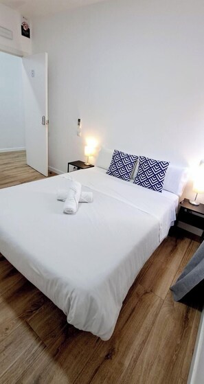 A neatly made bed with white linens and a rolled towel, two patterned pillows, a nightstand with a lamp, and a wooden floor.