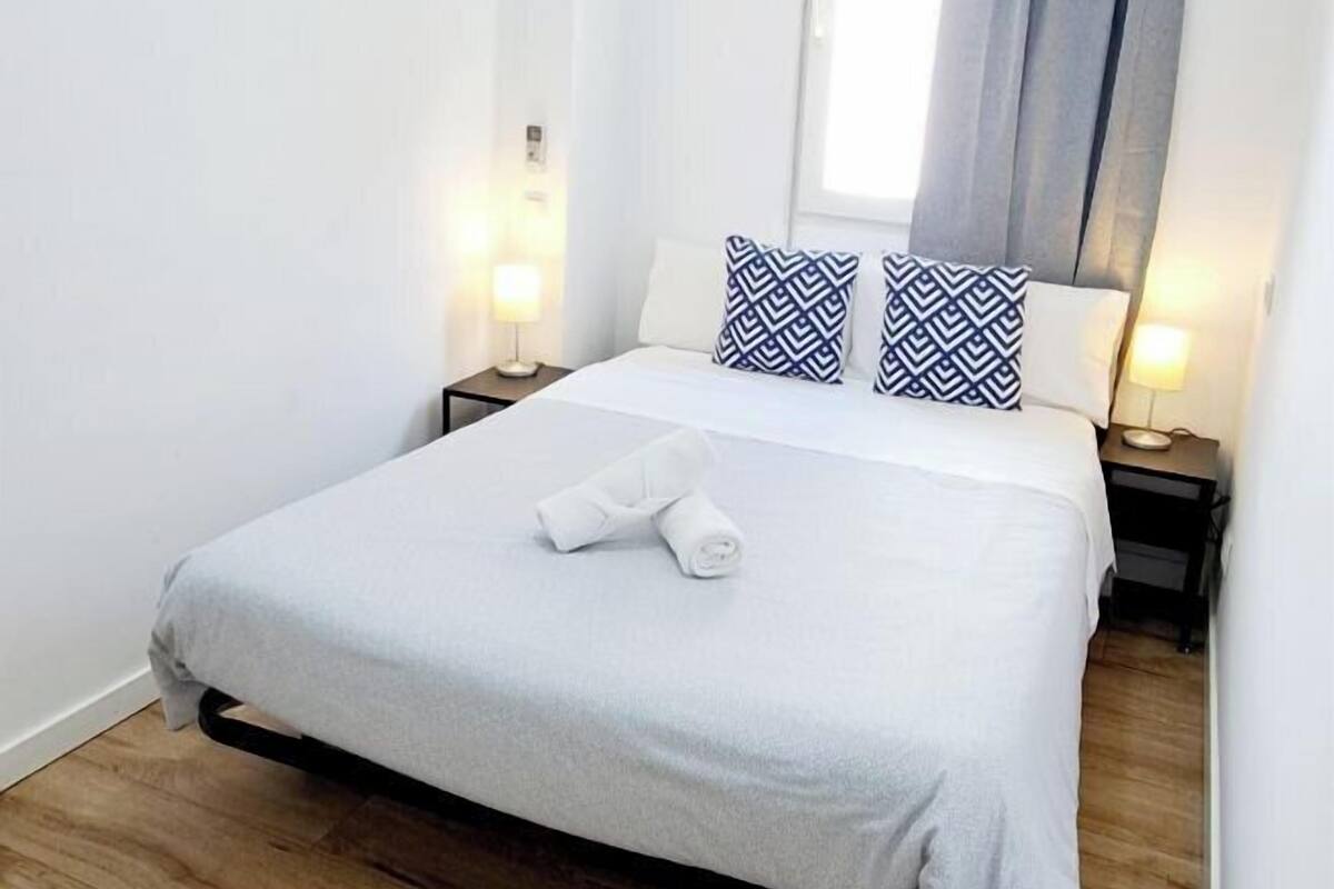 Un lit bien fait, avec une literie blanche et deux oreillers, une lampe de chevet et une fenêtre avec des rideaux.
