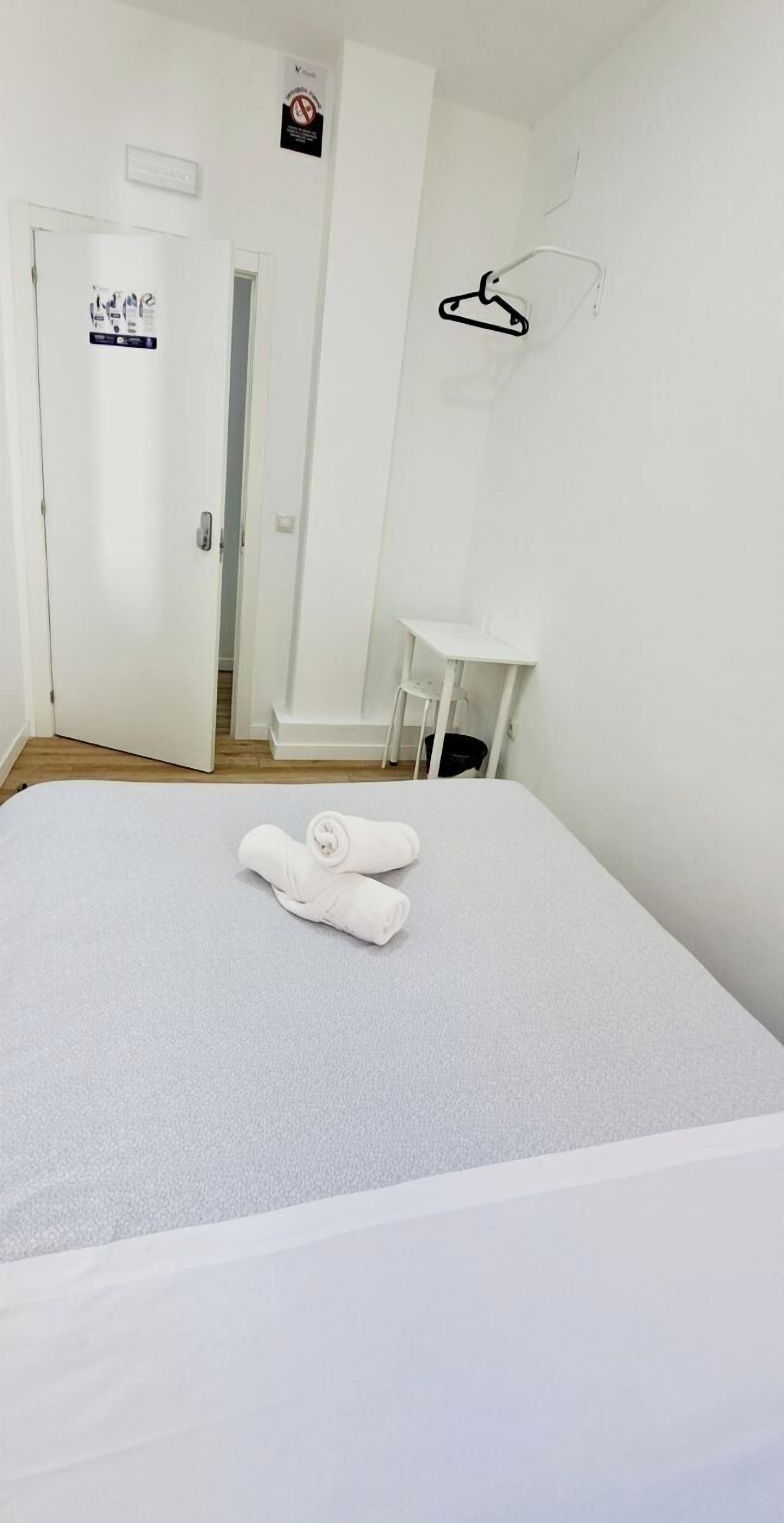 A hotel room with a bed, a white door, a white wall, a white chair, and a white table.