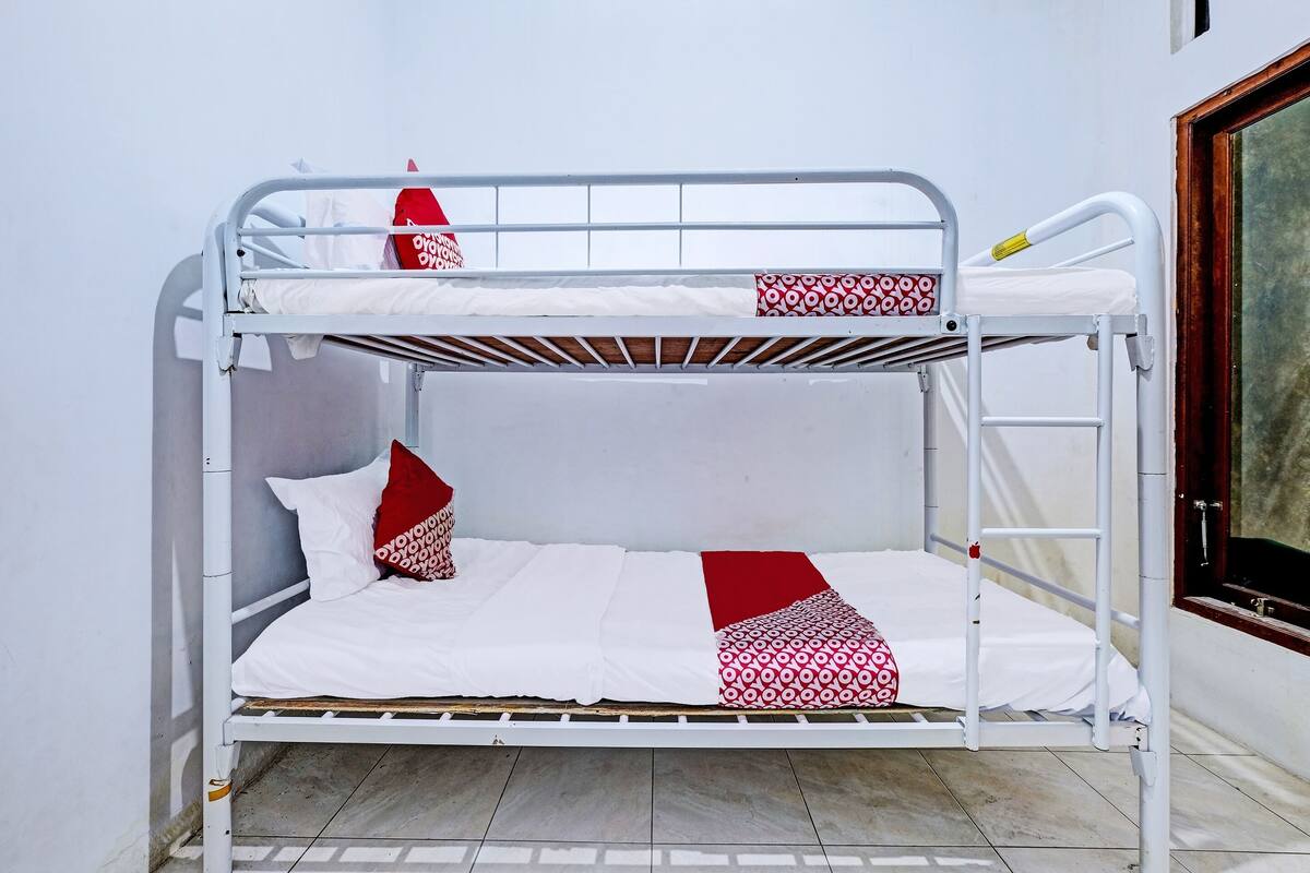 Una cama litera con ropa de cama blanca y detalles rojos, una ventana con vistas y una puerta a la derecha.