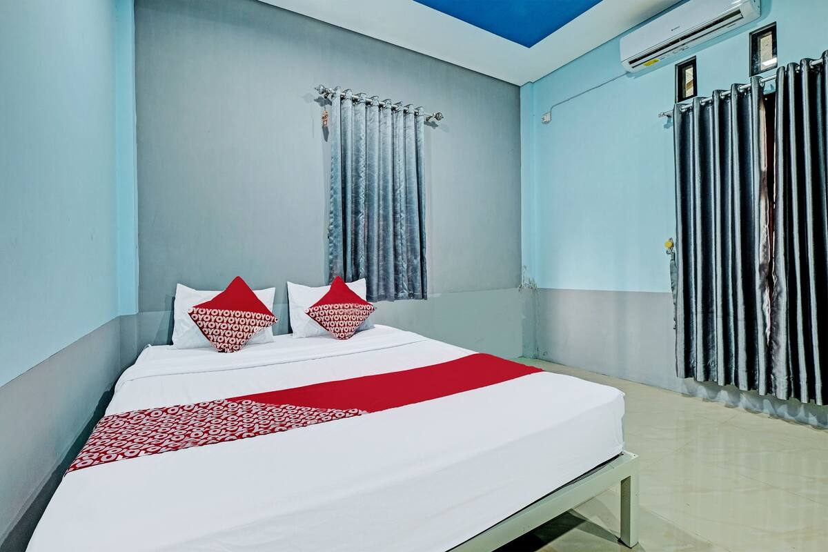 Una cama bien hecha con una colcha blanca y roja, dos almohadas y una ventana con cortinas.