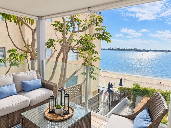 A balcony with a view of the beach, a wicker sofa, a glass table with candles, and a potted plant. - Bay-view 2BR condo in Crown Point with pool, patio, and beach access. (San Diego)