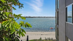 A view of a waterfront property with a building overlooking a calm body of water, a sandy beach, and a distant shoreline with buildings. - Bay-view 2BR condo in Crown Point with pool, patio, and beach access. (San Diego)