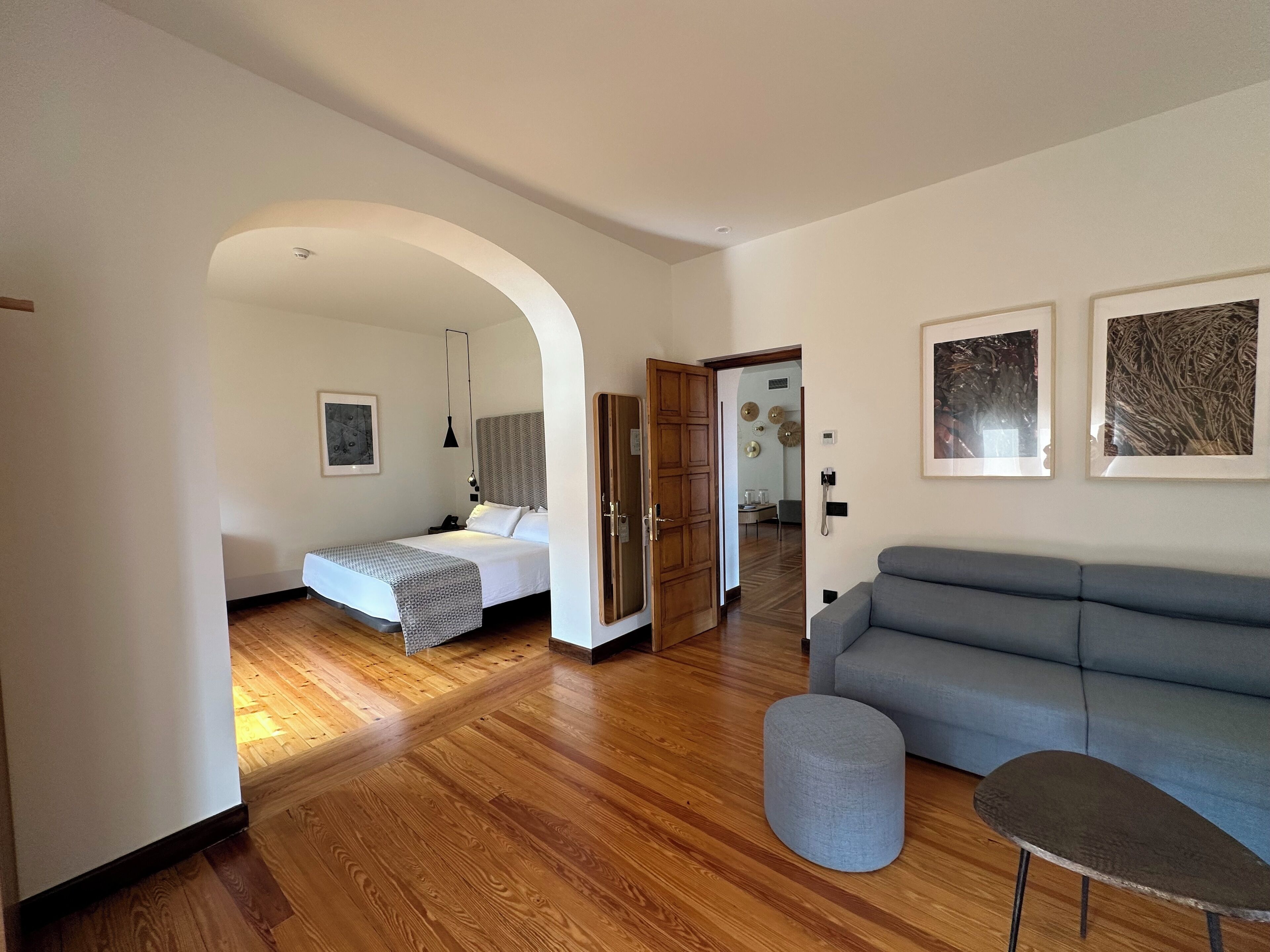 A modern living room with a grey sofa, a round ottoman, and a bed with a patterned bedspread.