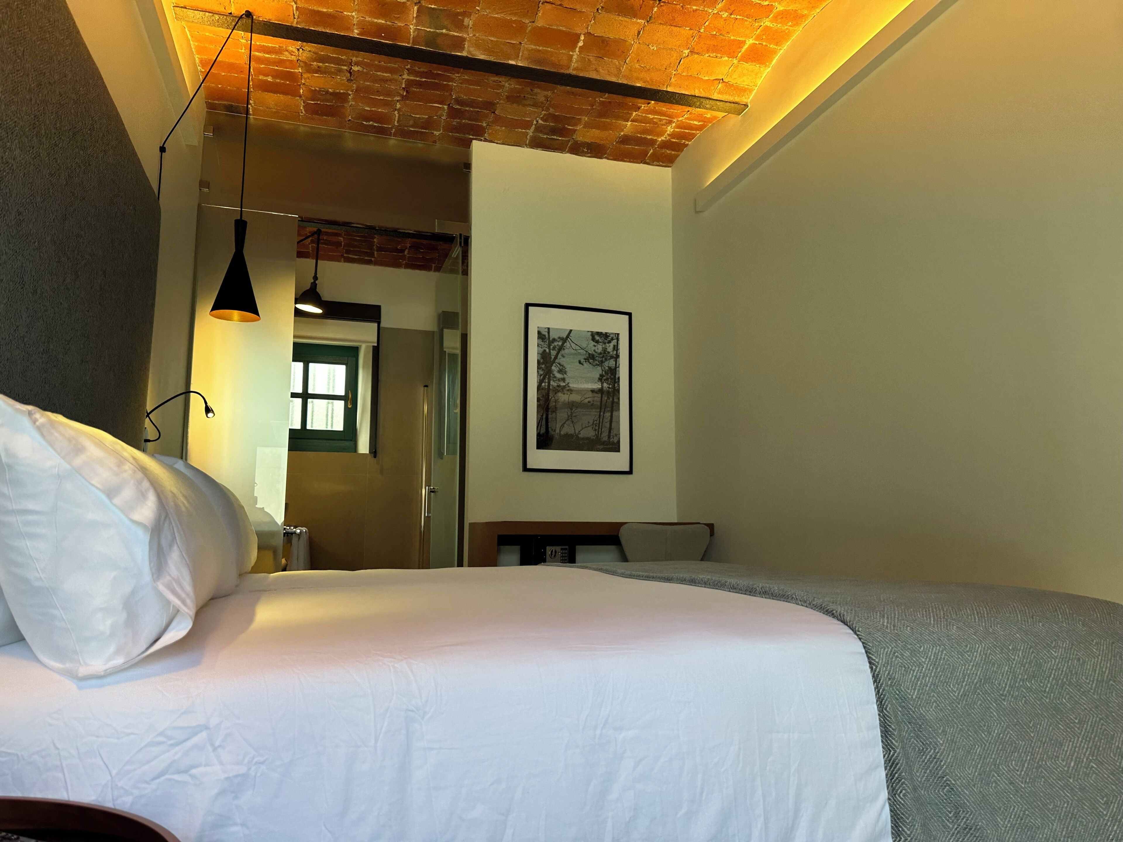 A bedroom with a bed, a desk, a chair, a framed picture on the wall, and a brick ceiling.