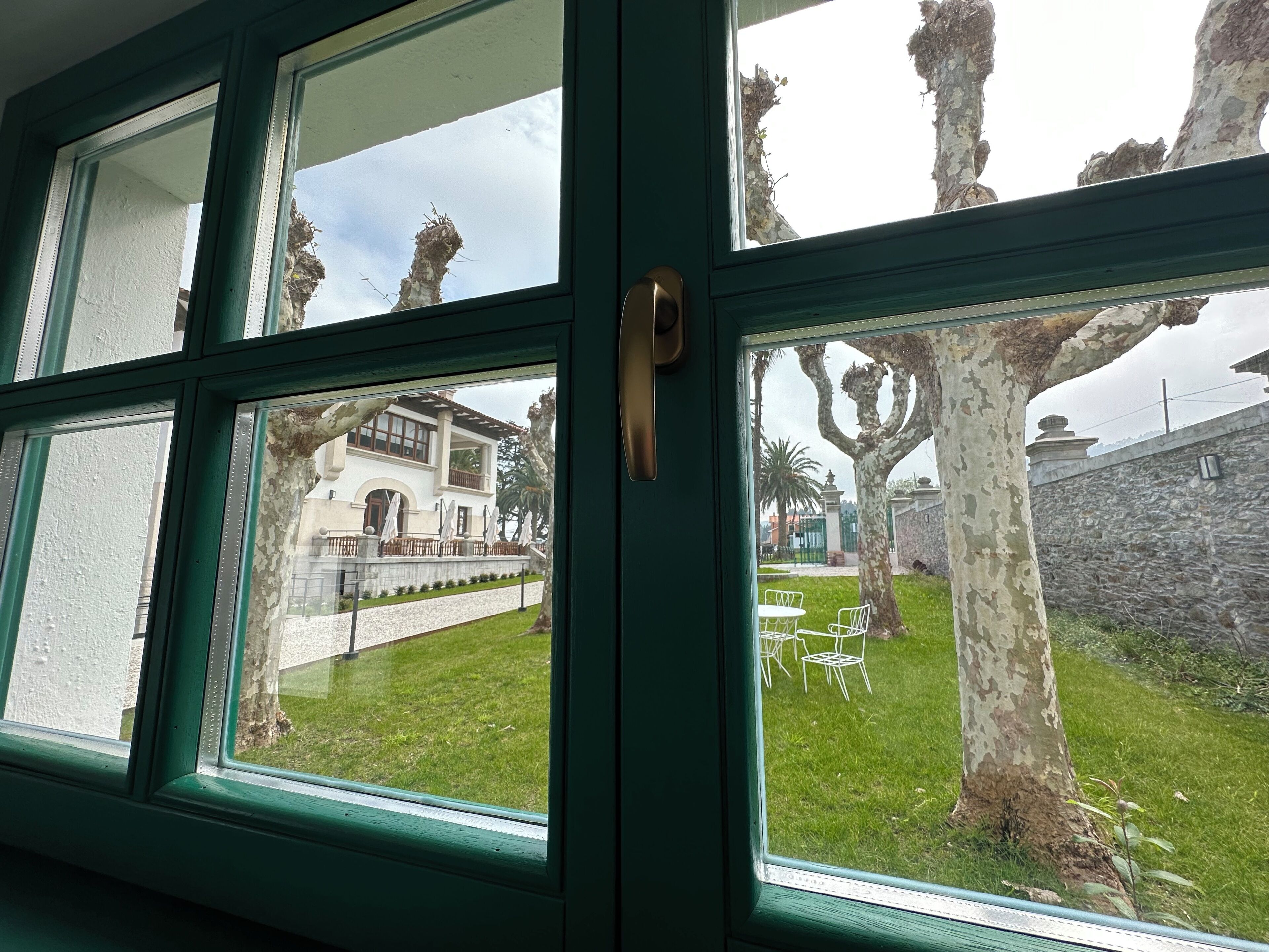 A view through a window with a green frame, revealing a white building with a stone wall and a grassy area with white chairs.