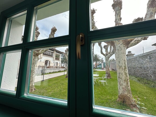 A view through a window with a green frame, revealing a white building with a stone wall and a grassy area with white chairs.