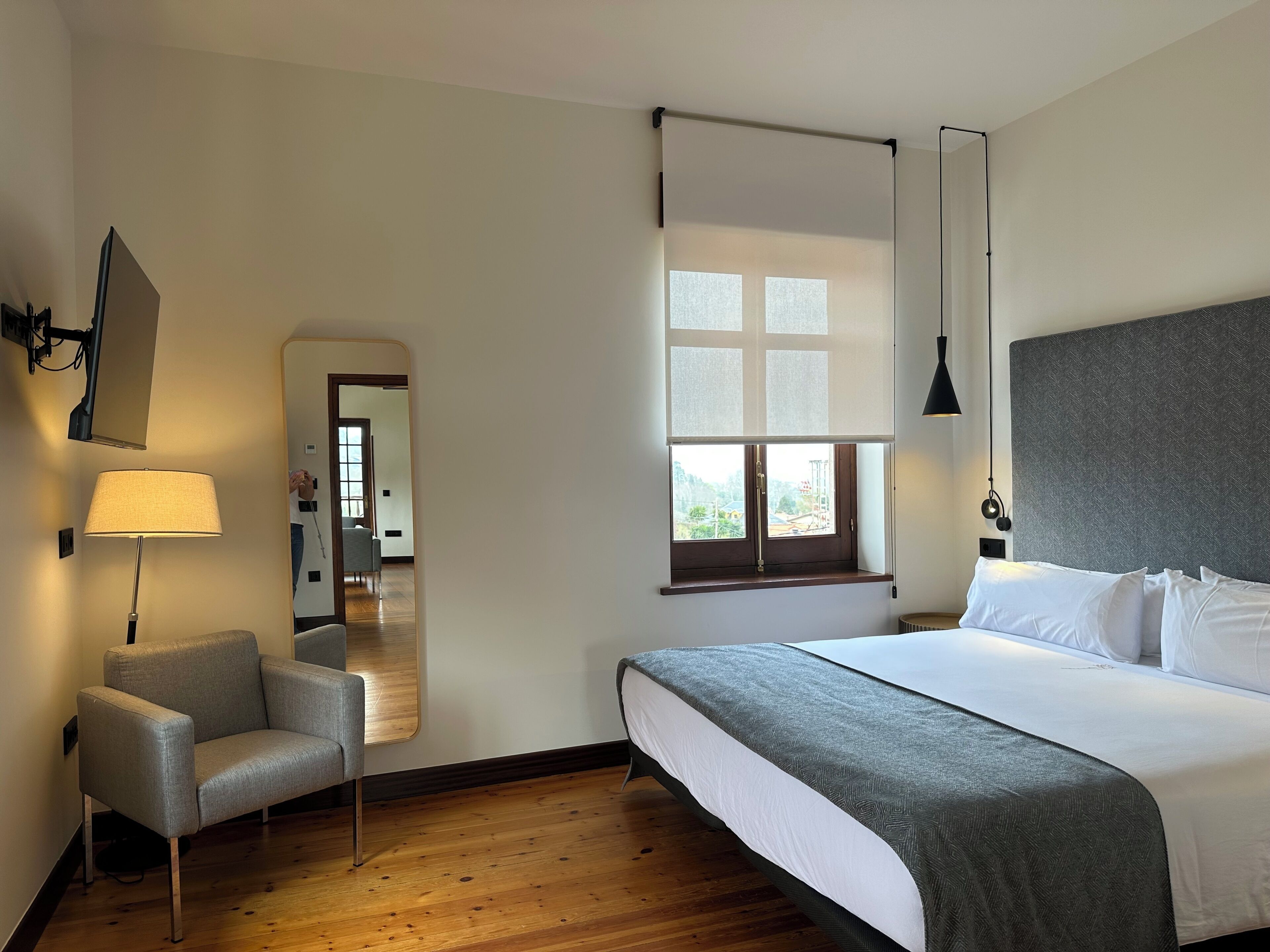A hotel room with a bed, a chair, a mirror, a window with blinds, and a lamp.