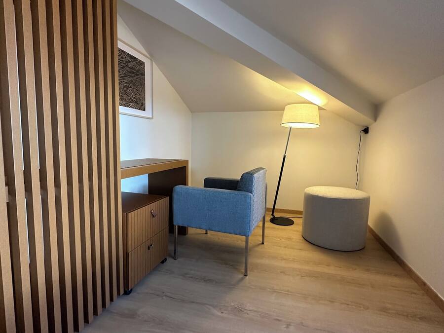Un dormitorio moderno en ático con techo inclinado, un separador de madera, una silla azul, un otoman redondo y una lámpara de pie.
