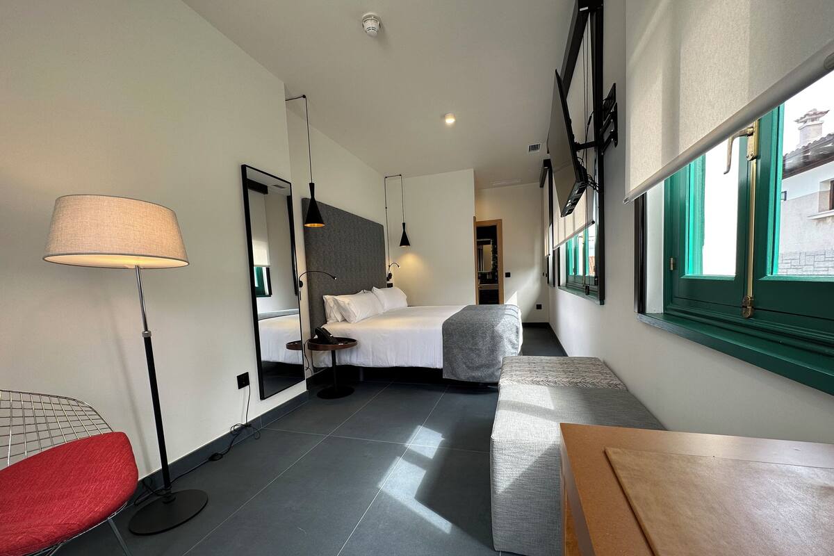 A modern hotel room with a bed, bedside table, lamp, and a view of the outside.