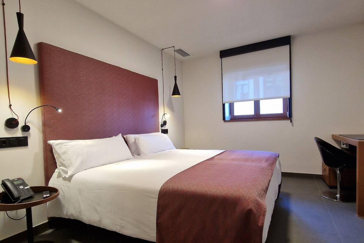 A hotel room with a bed, bedside table, lamp, and a desk with a chair.