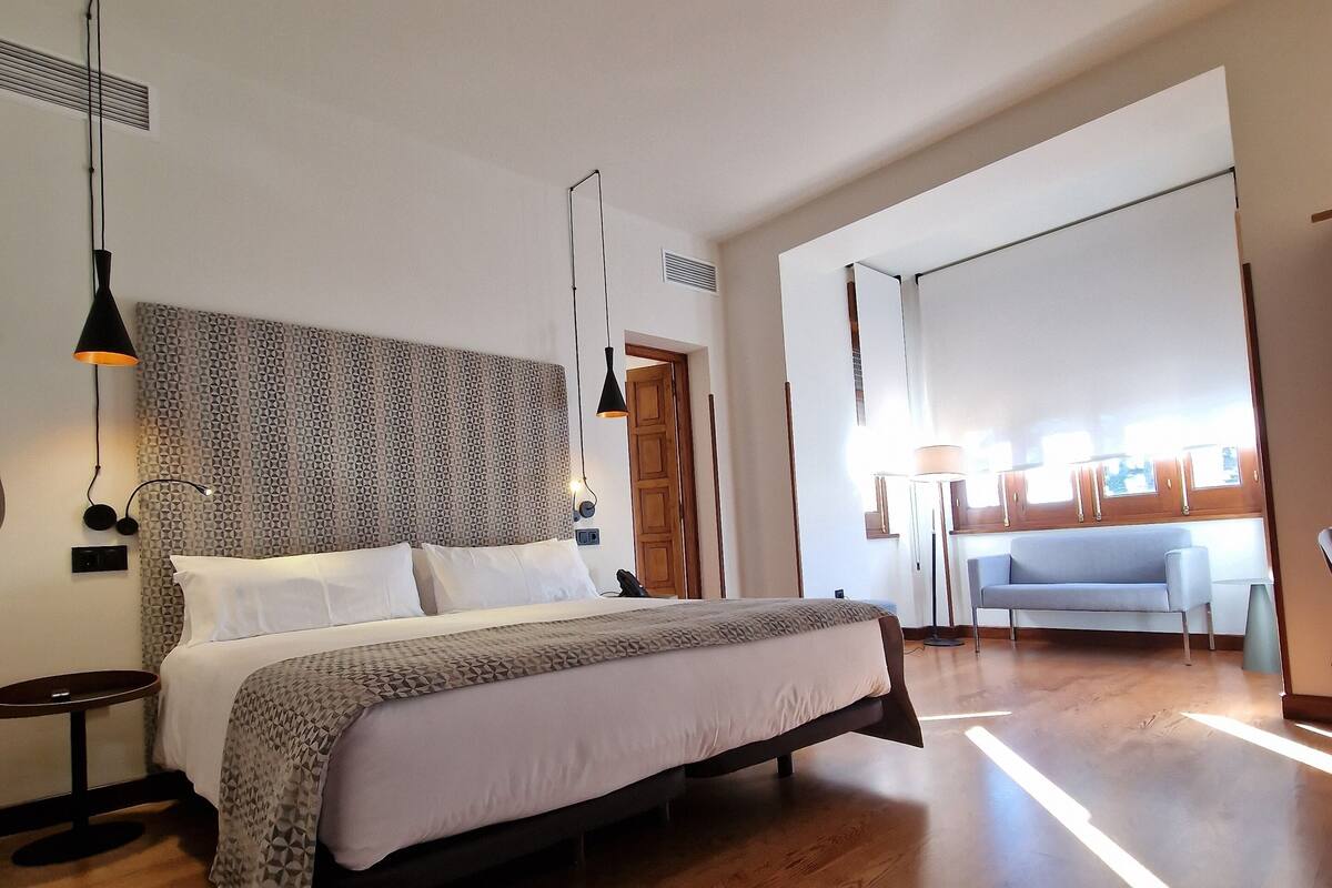 A modern hotel room with a large bed, wooden flooring, and a view of the city through a large window.