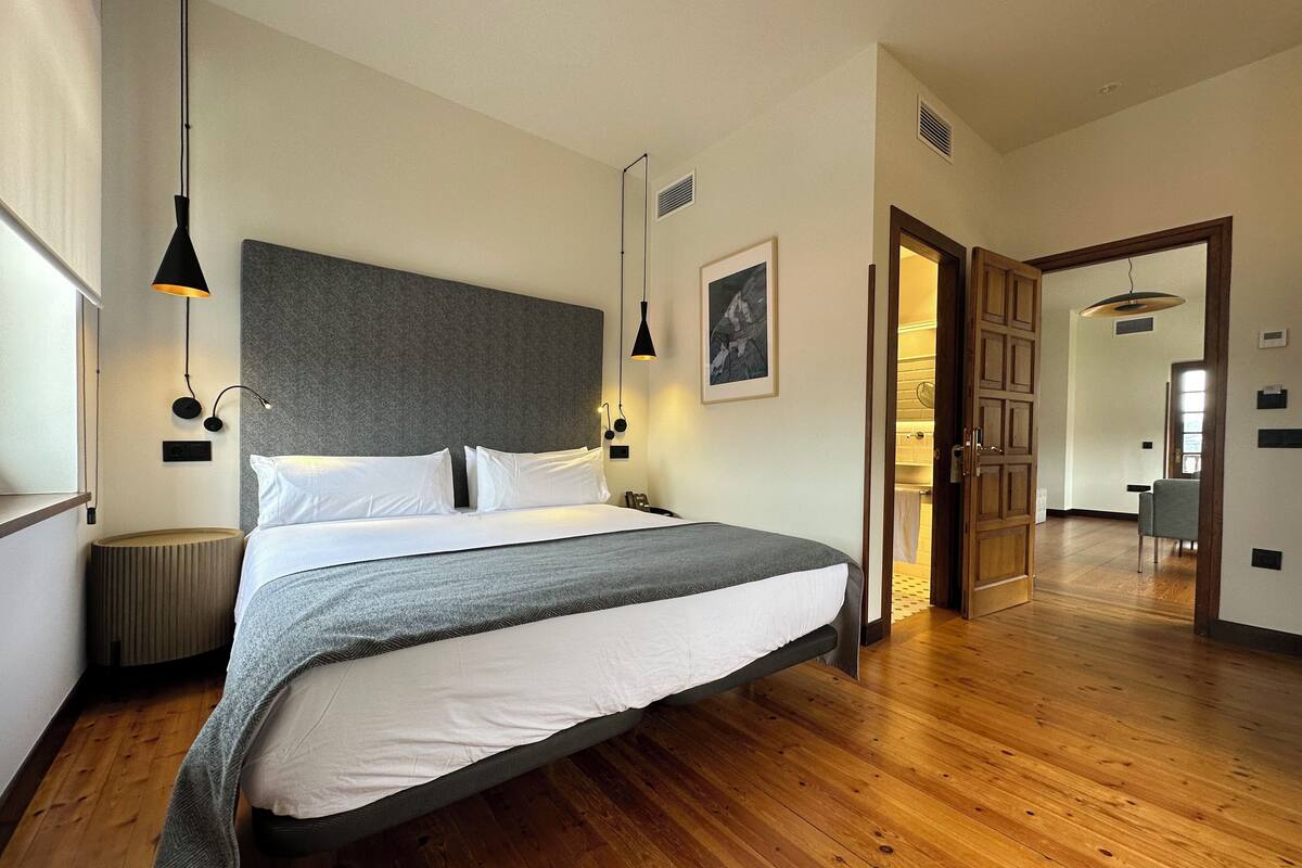 A modern bedroom with a large bed, wooden flooring, and a view into the bathroom.