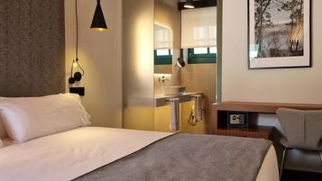 Habitación de hotel con cama, escritorio, silla, baño con espejo y un cuadro en la pared.