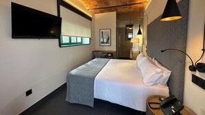 Una habitación de hotel moderna con techo de ladrillo, un televisor de pantalla plana montado en la pared, una cama con ropa blanca, una mesita de noche con lámpara y un teléfono.