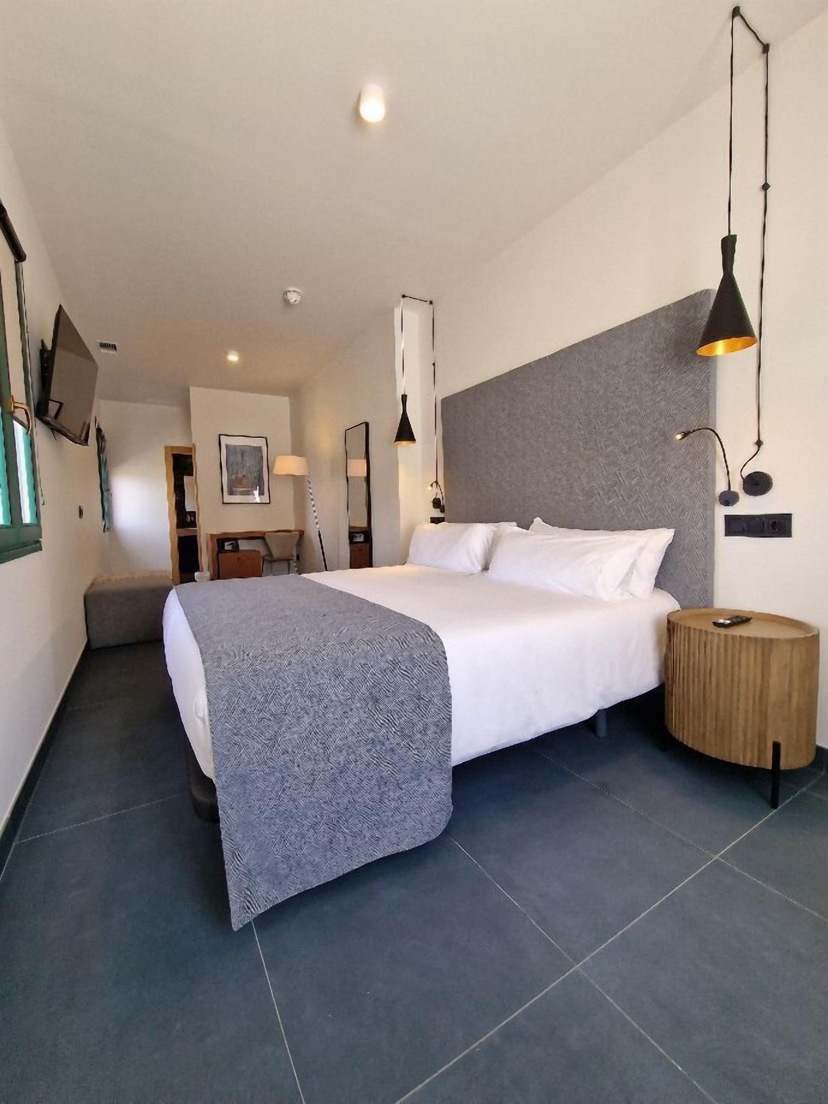 Une chambre d’hôtel moderne avec un grand lit, une petite table d’appoint ronde en bois et un plaid gris à texture intéressante.