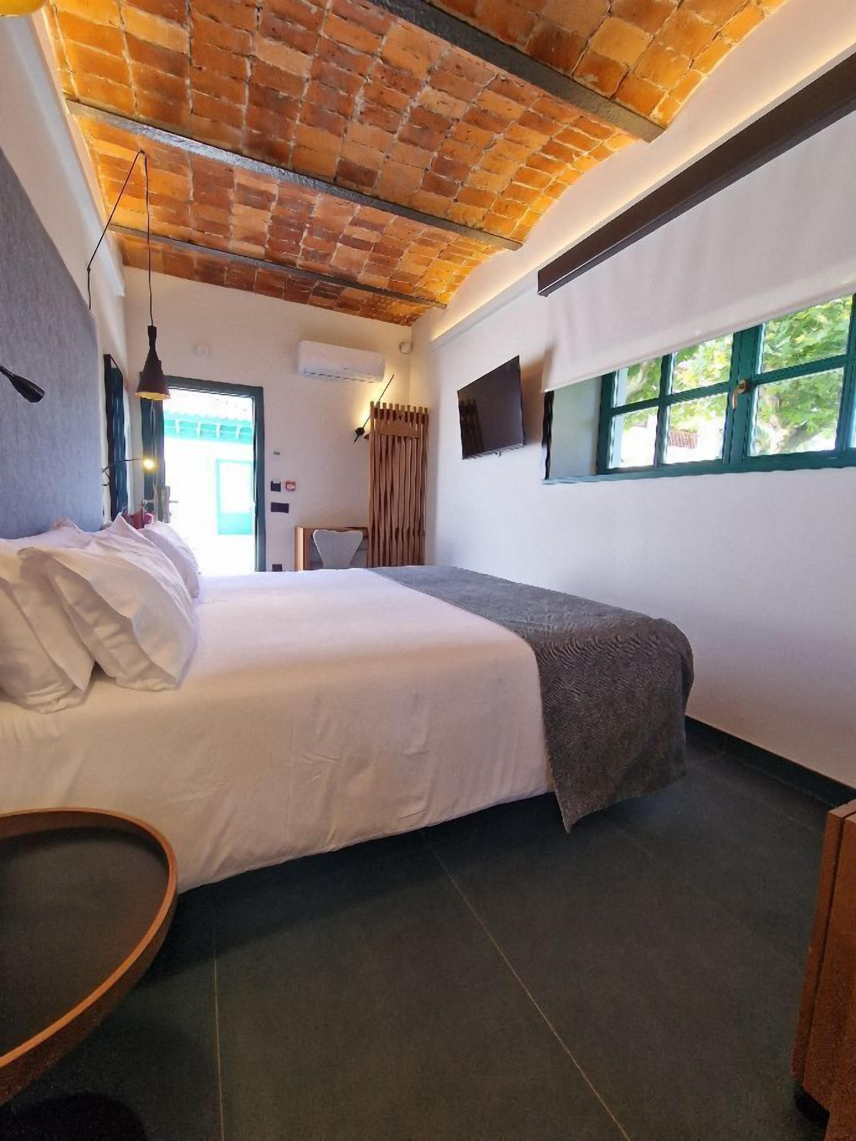 A bedroom with a bed, a TV mounted on the wall, a window, and a ceiling with exposed brick.