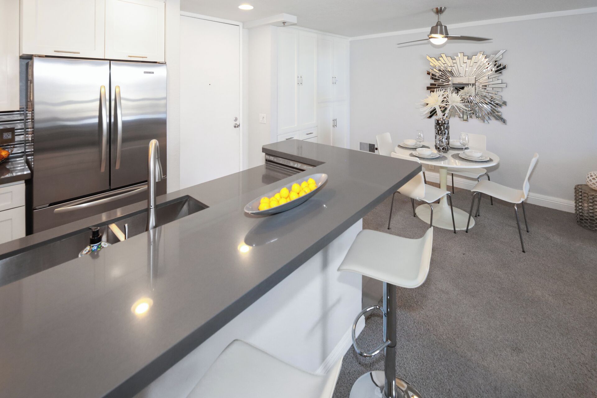 A modern kitchen with a stainless steel refrigerator, a grey countertop, and a dining area with white chairs and a table.