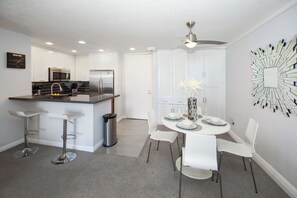 A modern kitchen with a central island, white cabinetry, and stainless steel appliances. There is a dining area with a round table, white chairs, and a decorative mirror on the wall.