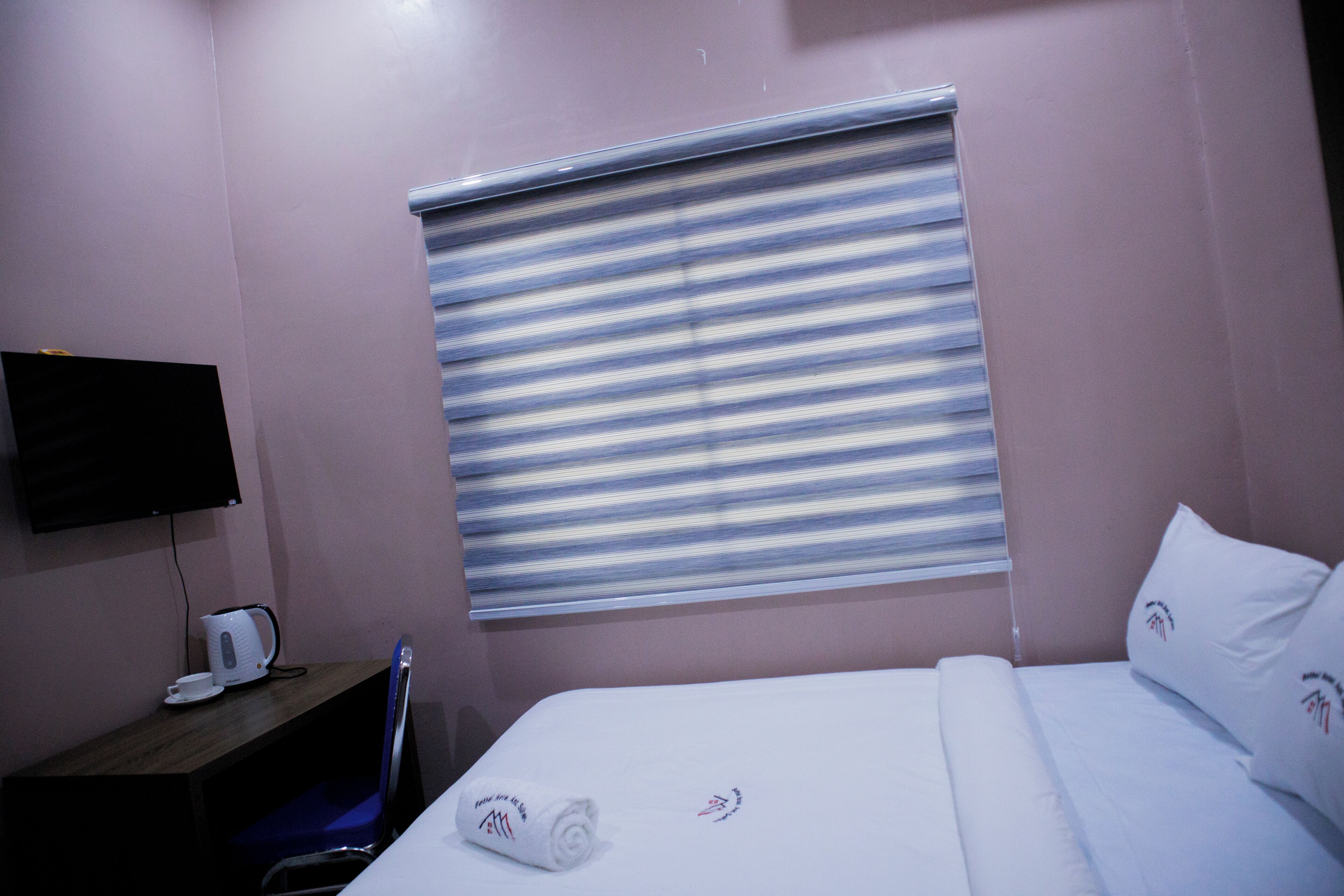 A hotel room with a bed, a desk with a kettle, a chair, and a TV on the wall.