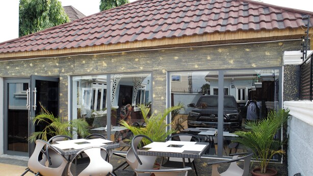 An outdoor cafe with tables and chairs, potted plants, and a building with a tiled roof. - Bethel Hotel and Suites (Abuja)