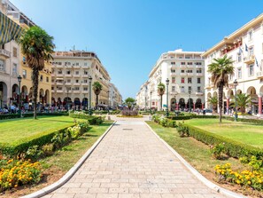 Une large promenade piétonne dallée, bordée d’immeubles dotés de balcons et de fenêtres en arc de cercle, qui mène à un espace central ouvert, agrémenté de verdure et de fleurs.