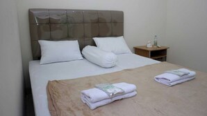 Habitación de hotel con una cama grande, dos almohadas, una mesita de noche con una botella y una caja de pañuelos, y dos toallas dobladas sobre la cama.