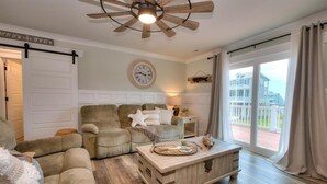 A cozy living room with a large window, a ceiling fan, and a clock on the wall. - Oceanview Hawks Landing in Oak Island: 3BR, 2BA retreat for 6 guests. (Oak Island)