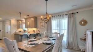 A dining area with a wooden table set for four, white chairs, pendant lighting, and a kitchen in the background. - Oceanview Hawks Landing in Oak Island: 3BR, 2BA retreat for 6 guests. (Oak Island)