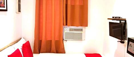 A hotel room with a bed, red pillows, a TV, and an air conditioning unit.