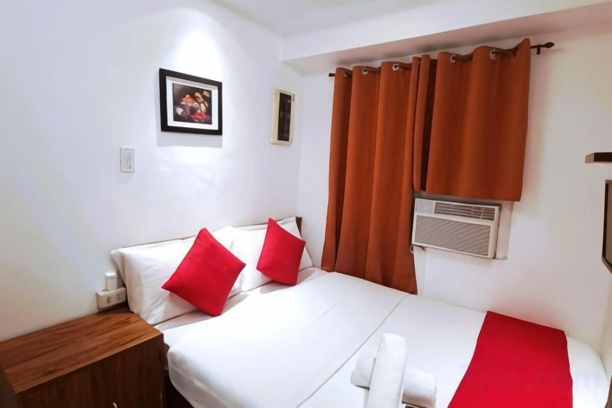 A single bed with red pillows, a bedside table, and a window with brown curtains.