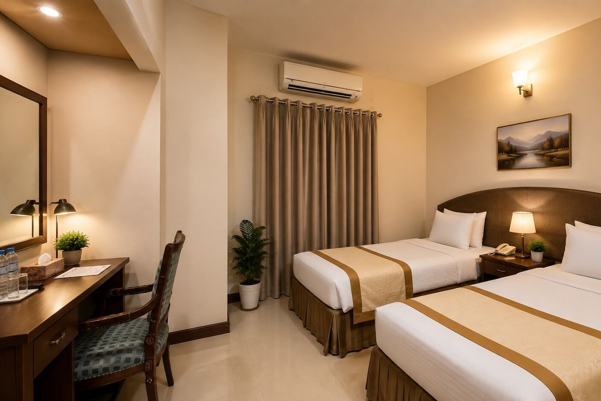 A hotel room with two beds, a desk with a chair, a desk lamp, a potted plant, an air conditioning unit, and a painting on the wall.