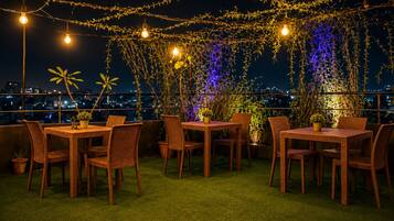 Un área de comedor al aire libre con mobiliario de mimbre y luces colgantes, con vista a un paisaje urbano.