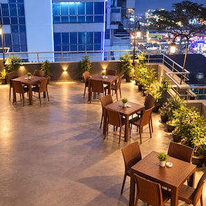 An outdoor dining area with tables and chairs, surrounded by potted plants, and overlooking a cityscape at night. - Hotel Fountain (Dhaka)