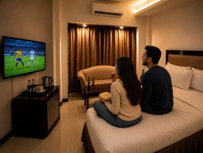 A hotel room with a television displaying a sports event, a sofa, a coffee table with snacks, and an air conditioning unit. - Hotel Horizon Blue (Dhaka)