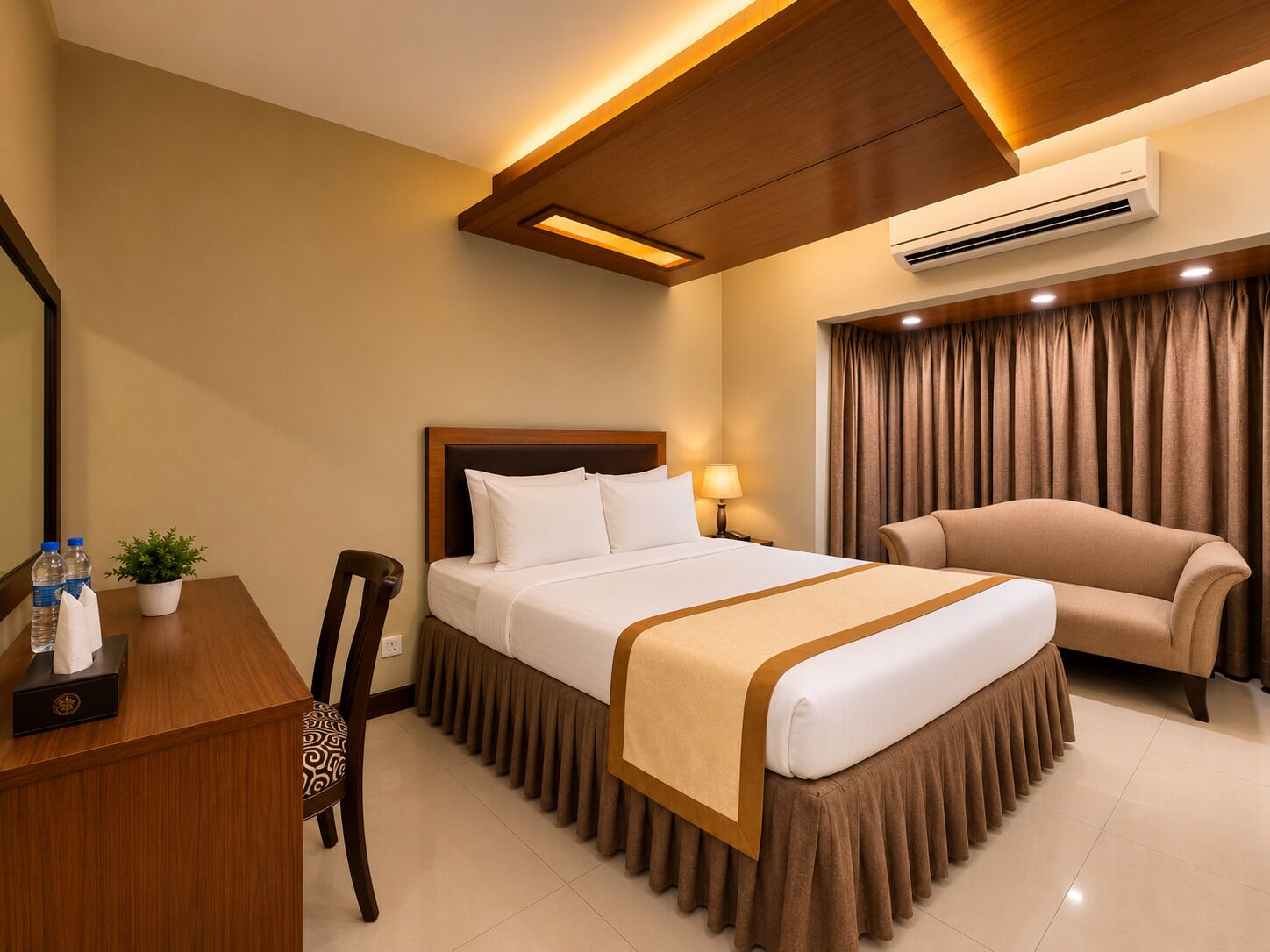 A hotel room with a bed, a desk with a chair, a sofa, an air conditioner, and a potted plant.
