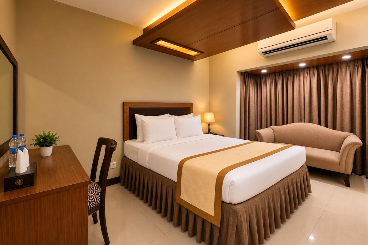 A hotel room with a bed, a desk with a chair, a sofa, an air conditioner, and a potted plant.